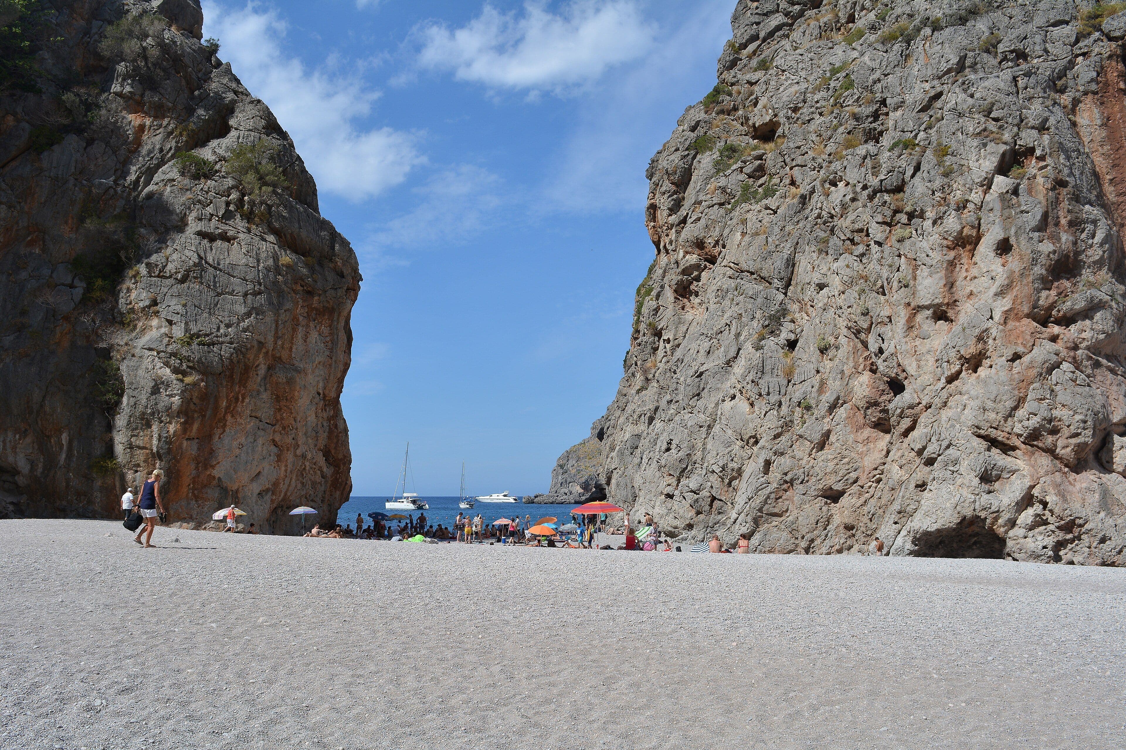 Platja de còdols blancs de sa Calobra encaixada entre dues grans parets de roca, amb velers fondejats al mar al fons