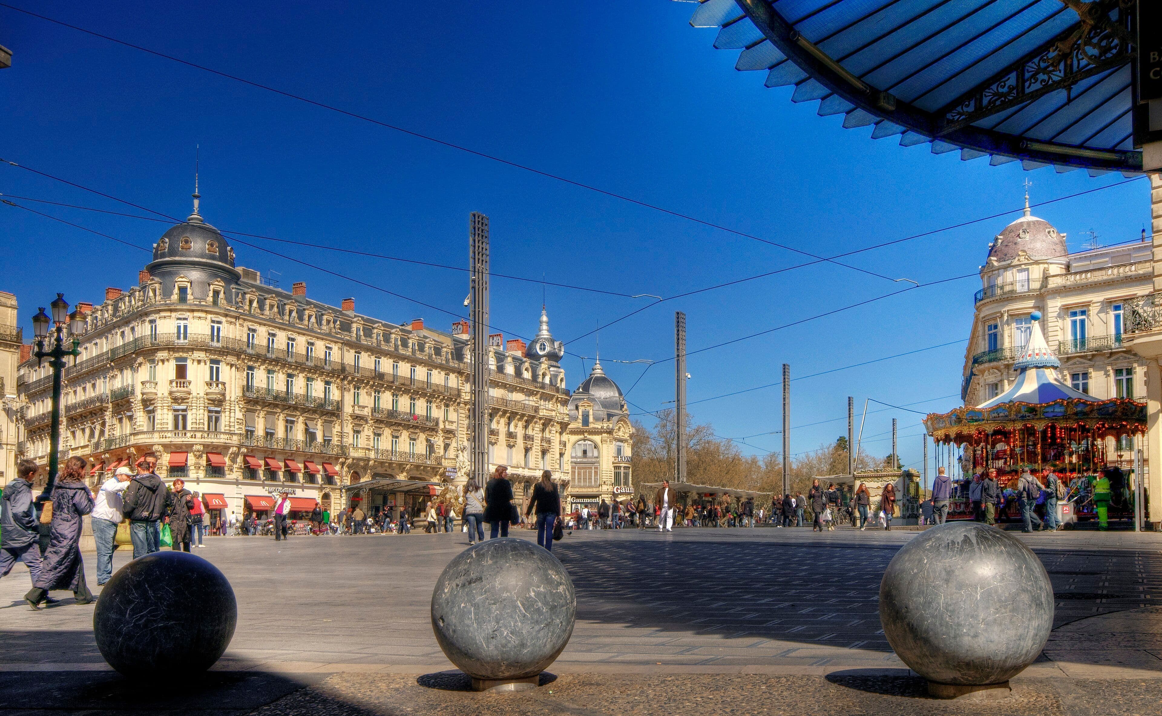 La plaza de la Comédie de Montpellier con sus edificios de piedra clara, el tiovivo y gente cruzando la explanada